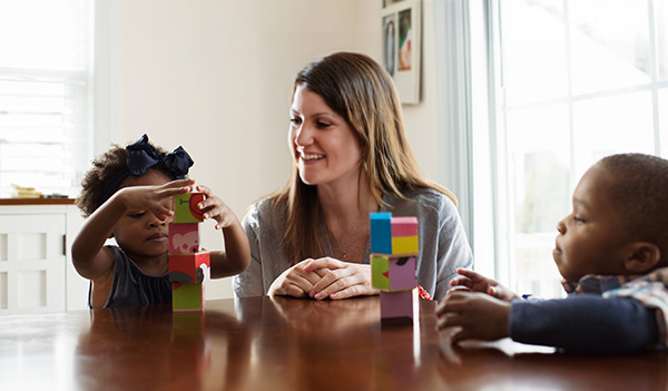 Female child care professional sitting at table with two children playing blocks.